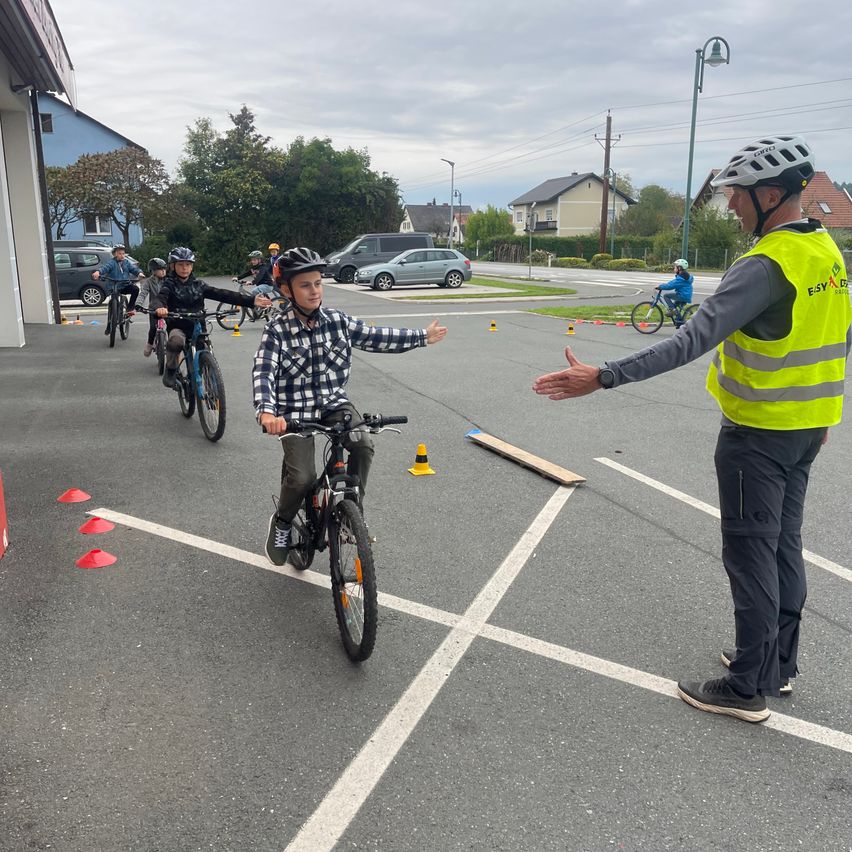 Eine Gruppe von Kindern auf Fahrrädern fährt auf einem Parkplatz. Sie tragen Helme. Ein Mann in einer reflektierenden Weste gibt Anweisungen.