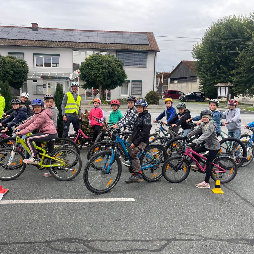 Eine Gruppe von Kindern mit Helmen fährt auf einer Straße mit Fahrrädern. Ein Mann in einer reflektierenden Weste steht hinter ihnen. Dahinter befinden sich Gebäude mit Solarpanelen.