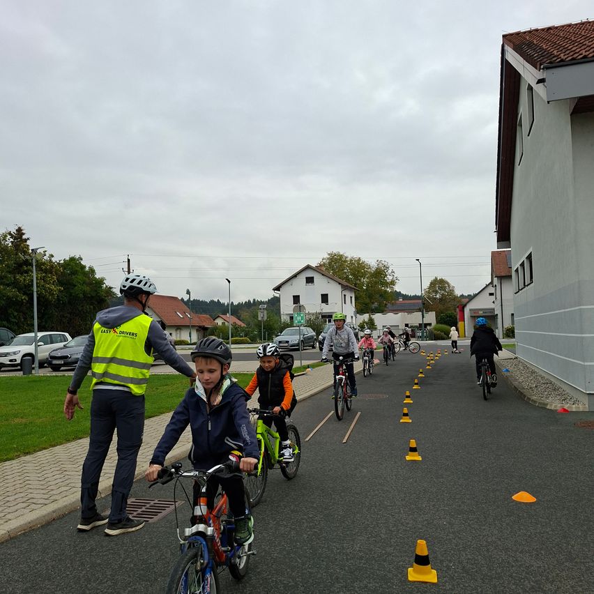Kinder fahren auf einer mit Hütchen gesäumten Straße mit Fahrrädern, wobei ein Erwachsener sie führt. Die Straße wird von Häusern und einem Rasenbereich flankiert.