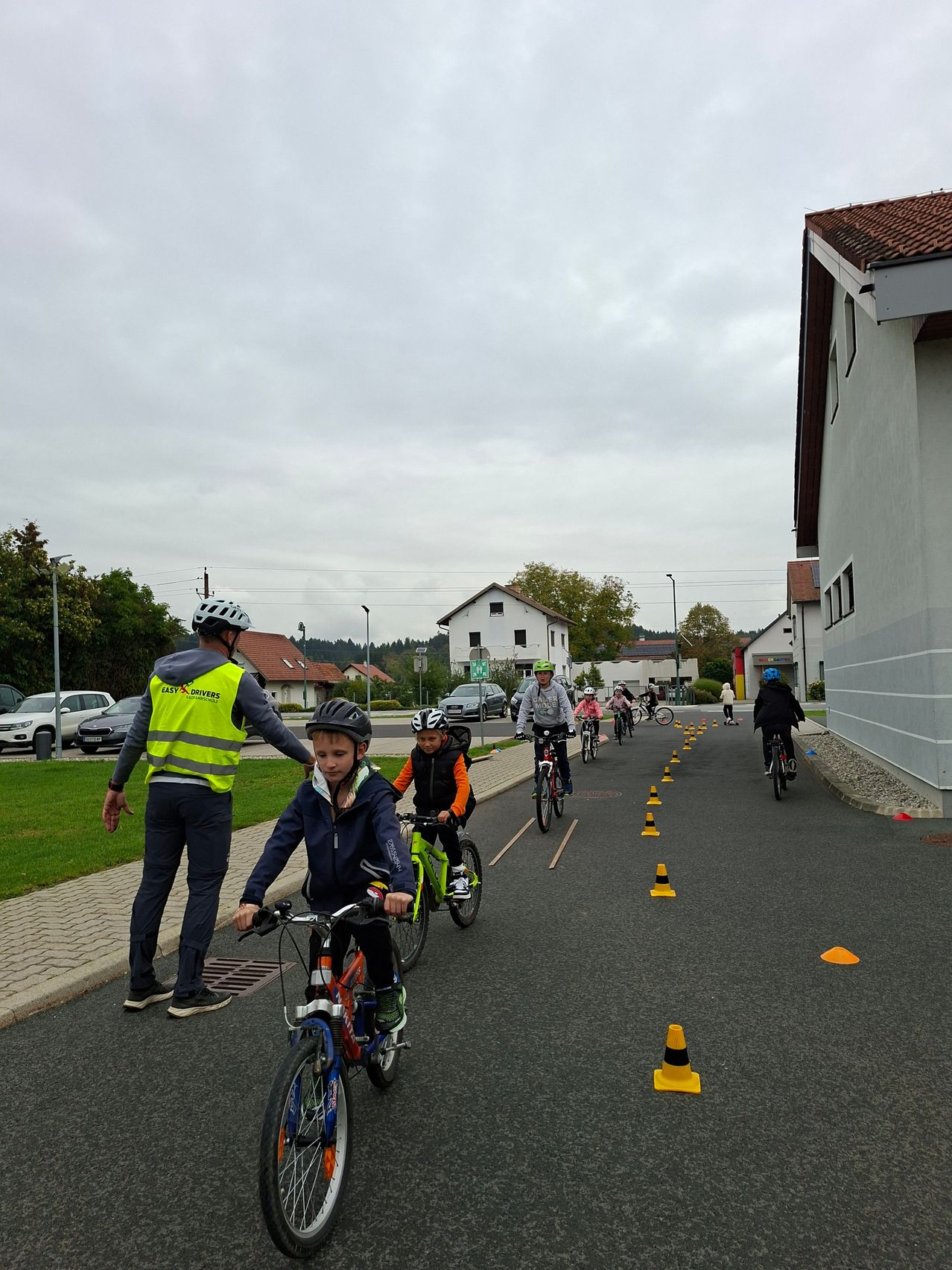 Kinder fahren auf einer mit Hütchen gesäumten Straße mit Fahrrädern, wobei ein Erwachsener sie führt. Die Straße wird von Häusern und einem Rasenbereich flankiert.