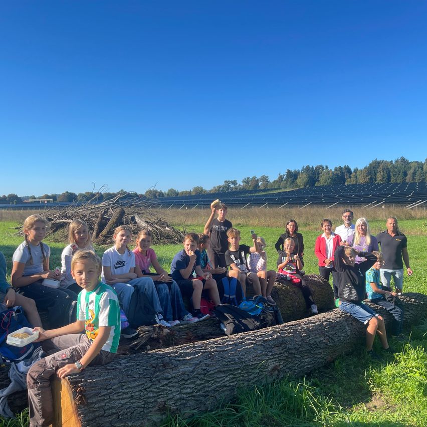 Eine Gruppe von Menschen, einschließlich Kindern, sitzt auf einem Baumstamm in einem Grasfeld mit einer Solaranlage im Hintergrund. Einige essen und machen Fotos.