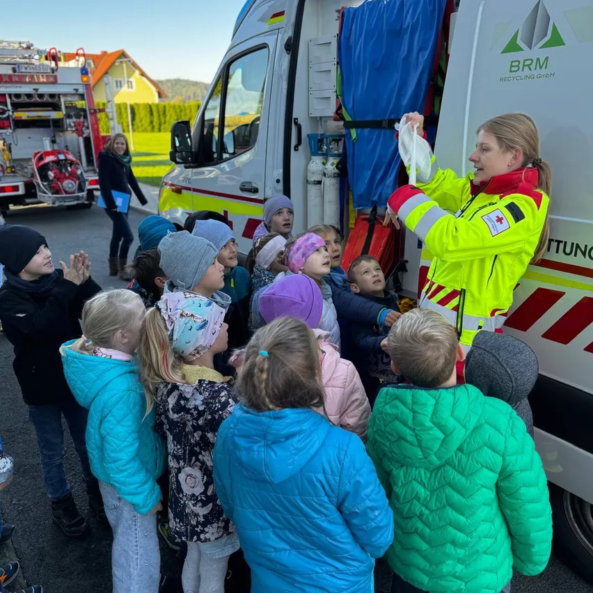 Eine Frau in einer gelben Sicherheitsjacke erklärt etwas einer Gruppe von Kindern vor einem Krankenwagen. Im Hintergrund ist ein Feuerwehrauto geparkt.