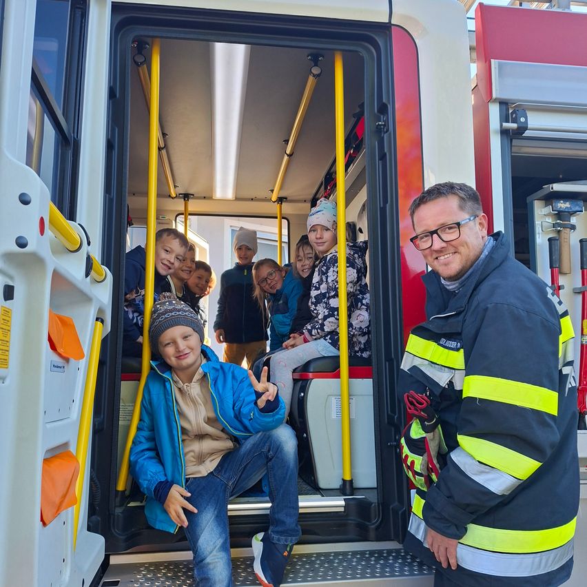 Eine Gruppe von Kindern und ein Feuerwehrmann sind in einem Krankenwagen und posieren lächelnd für ein Foto. Der Feuerwehrmann trägt eine reflektierende Weste.