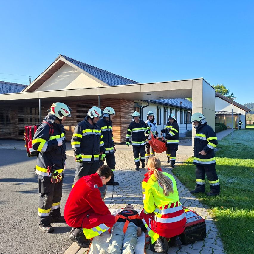 Eine Gruppe von Feuerwehrleuten und Sanitätern führt eine Trainingsübung vor einem Gebäude durch. Ein Dummy liegt auf dem Boden, betreut von zwei Personen in reflektierenden Westen.