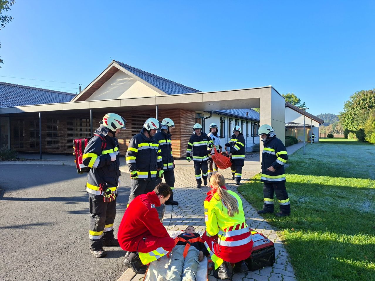 Eine Gruppe von Feuerwehrleuten und Sanitätern führt eine Trainingsübung vor einem Gebäude durch. Ein Dummy liegt auf dem Boden, betreut von zwei Personen in reflektierenden Westen.