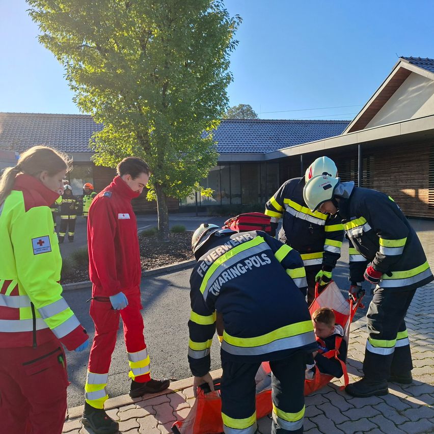 Feuerwehrleute in Uniformen bereiten sich darauf vor, ein Kind auf einer Trage vor einem Gebäude an einem sonnigen Tag zu transportieren.