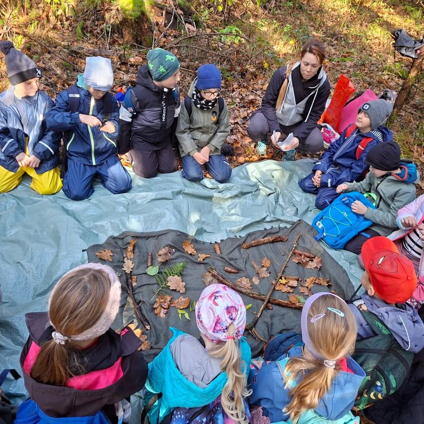 Eine Gruppe von Kindern und eine erwachsene Frau sitzen in einem Kreis auf einer Plane im Wald und untersuchen Blätter und Zweige. Sie sind warm gekleidet in Jacken und Mützen. Die Frau hält einen kleinen Behälter.
