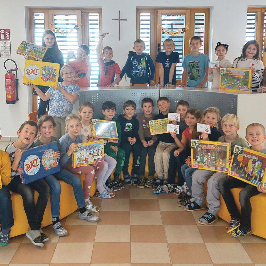 Gruppe von Kindern, die verschiedene Brettspiele und Bücher halten. Sie sitzen auf einer gelben Couch und stehen lächelnd in einem Klassenzimmer. Einige Kinder halten Urkunden. Hinter ihnen steht ein Tisch mit einem Kreuzsymbol.