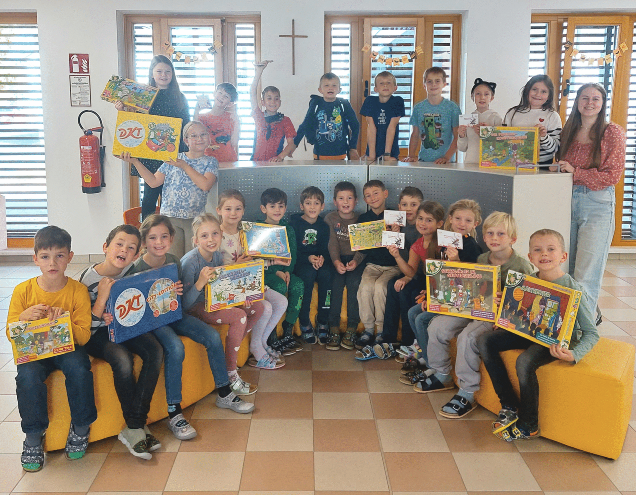 Gruppe von Kindern, die verschiedene Brettspiele und Bücher halten. Sie sitzen auf einer gelben Couch und stehen lächelnd in einem Klassenzimmer. Einige Kinder halten Urkunden. Hinter ihnen steht ein Tisch mit einem Kreuzsymbol.