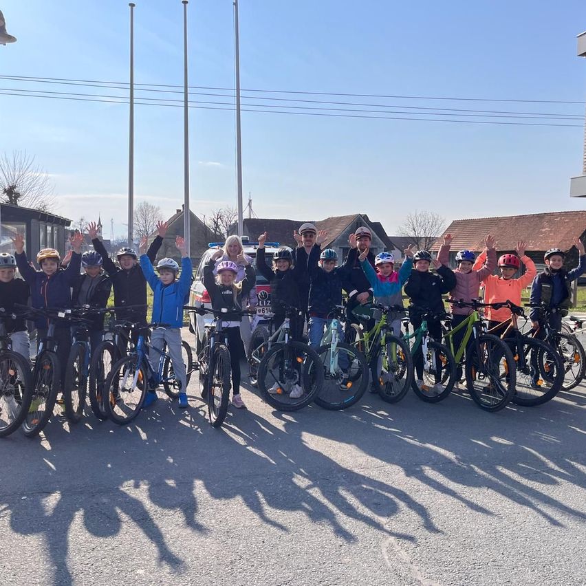 Eine Gruppe von Kindern und Erwachsenen steht mit ihren Fahrrädern in einem Außenbereich unter klarem Himmel. Einige tragen Helme, und im Hintergrund befinden sich Häuser und Versorgungsmasten.