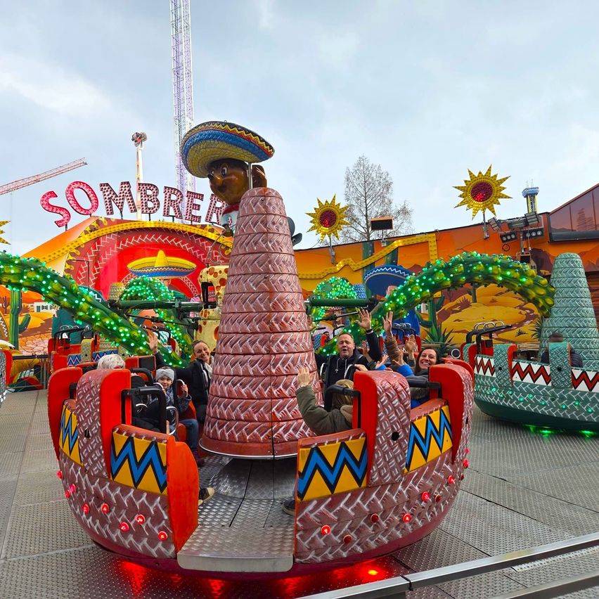 Eine Gruppe von Menschen fährt eine farbenfrohe, sombrero-thematische Achterbahn mit bunten Lichtern auf einem Jahrmarkt.