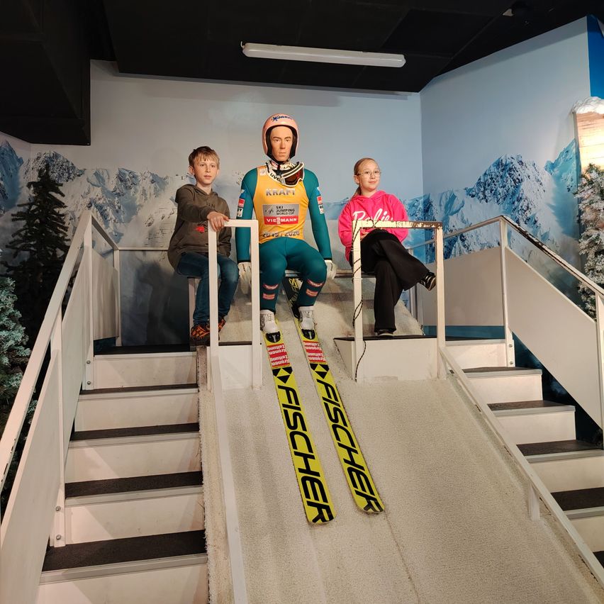 Drei Personen auf einem Skilift, eine in einem Skianzug, eine weitere in einer rosa Jacke und die dritte in einer braunen Jacke. Eine Wand im Hintergrund zeigt Berge.