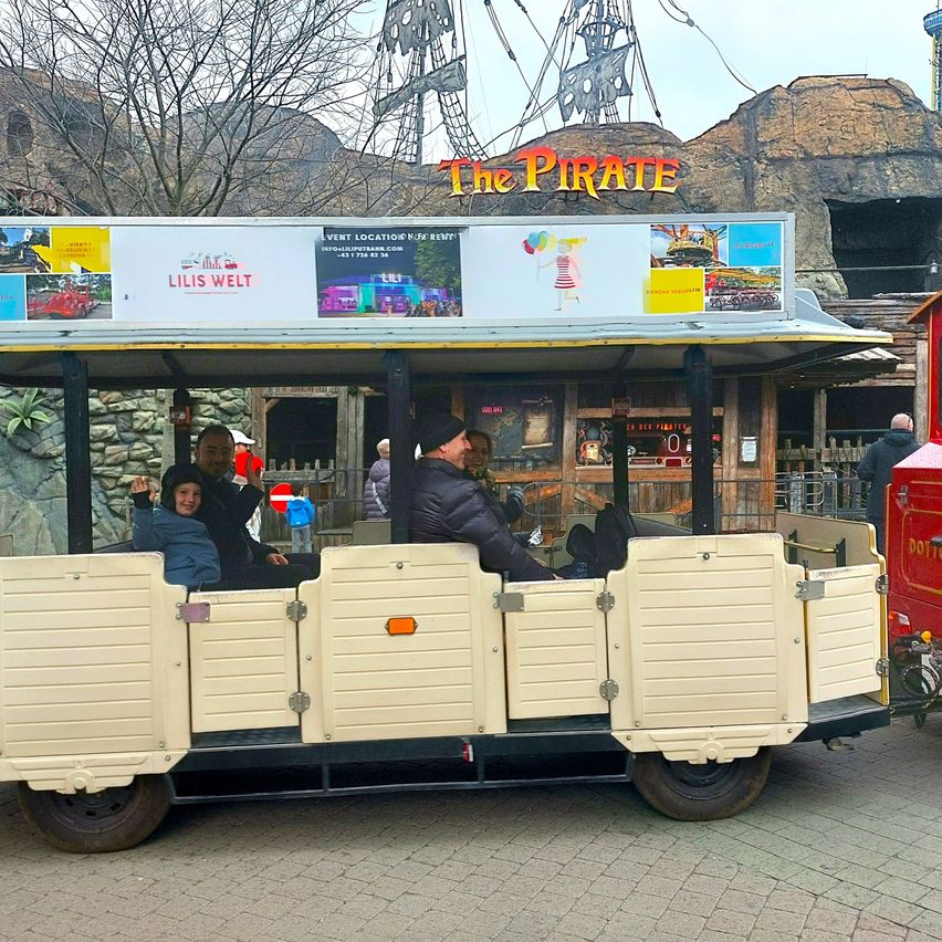 Eine Familie sitzt in einer Achterbahn im Vergnügungspark The Pirate. Die Achterbahn hat eine Werbung auf dem Dach. Leute stehen um die Achterbahn herum.