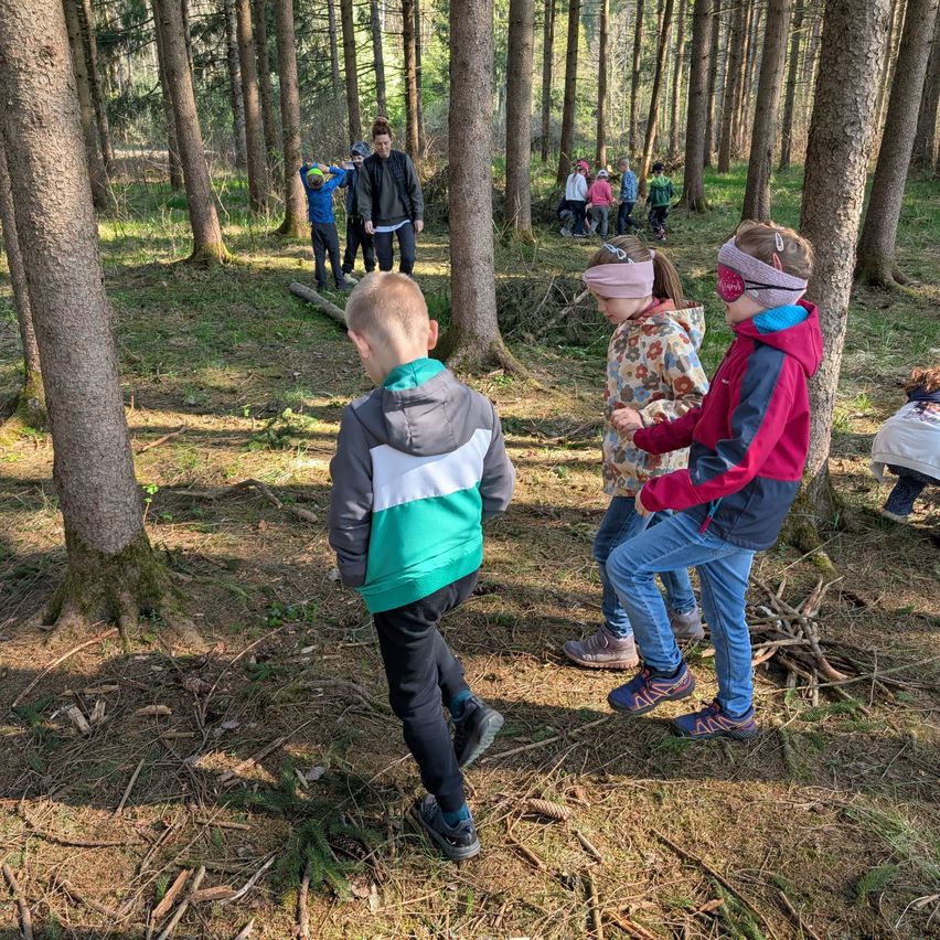 Kinder in Jacken und Mützen spielen im Wald, einige mit verbundenen Augen. Hohe Bäume umgeben sie, im Hintergrund sind weitere Kinder zu sehen.