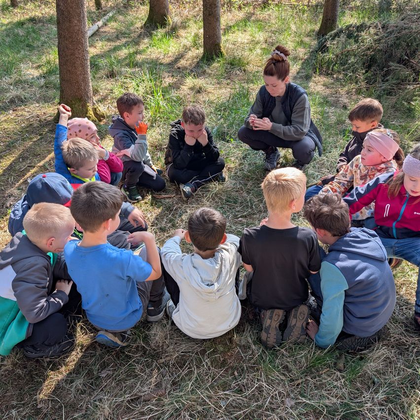 Eine Gruppe von Kindern sitzt im Kreis auf einem Grasfeld mit einer erwachsenen Frau, die vor ihnen kniet. Die Kinder tragen Jacken und Turnschuhe.