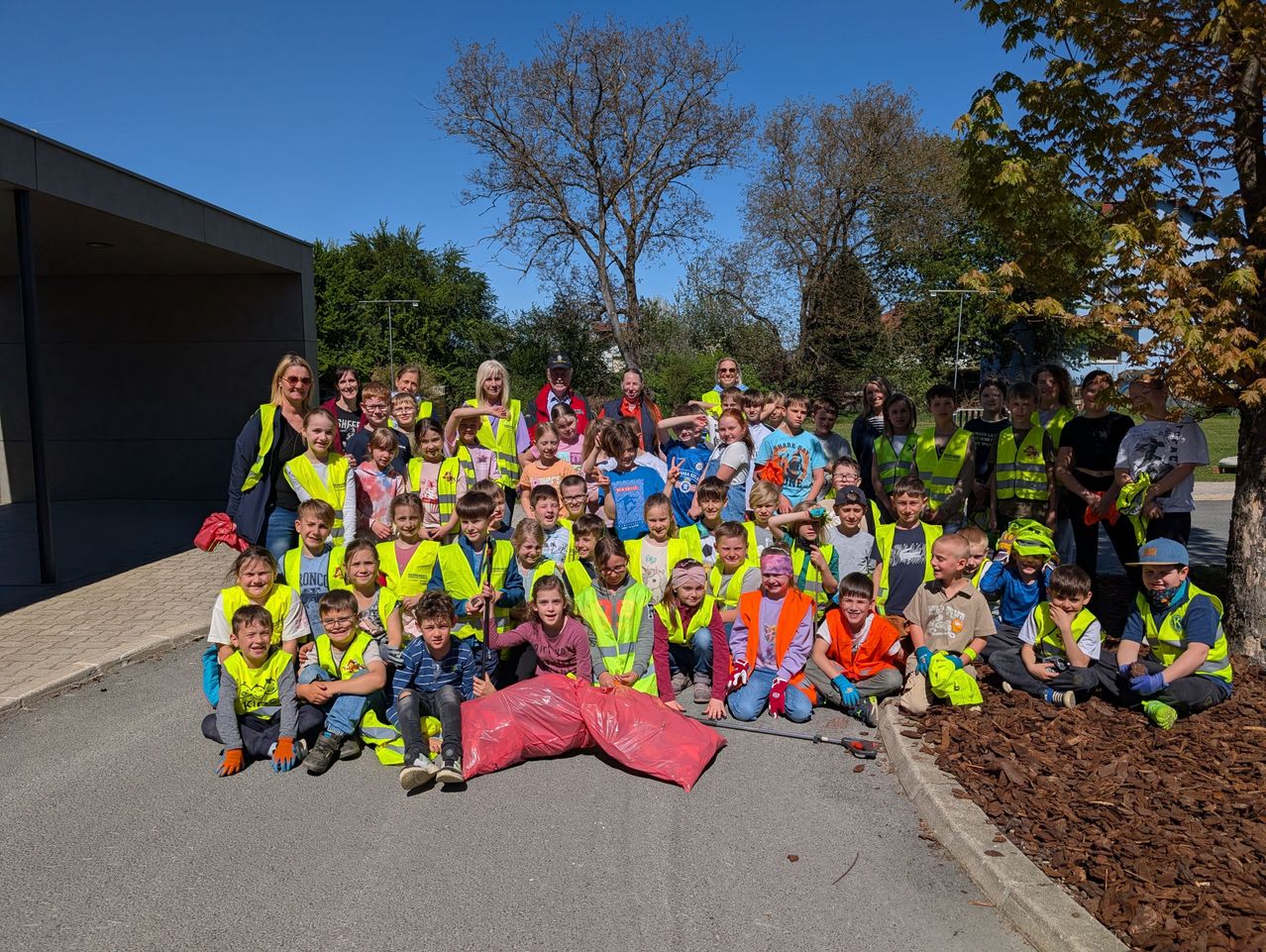 Eine Gruppe von Kindern und Erwachsenen posiert für ein Foto in einem Park, alle tragen gelbe Westen und halten Plastiktüten. Einige haben Gesichtsbemalung.
