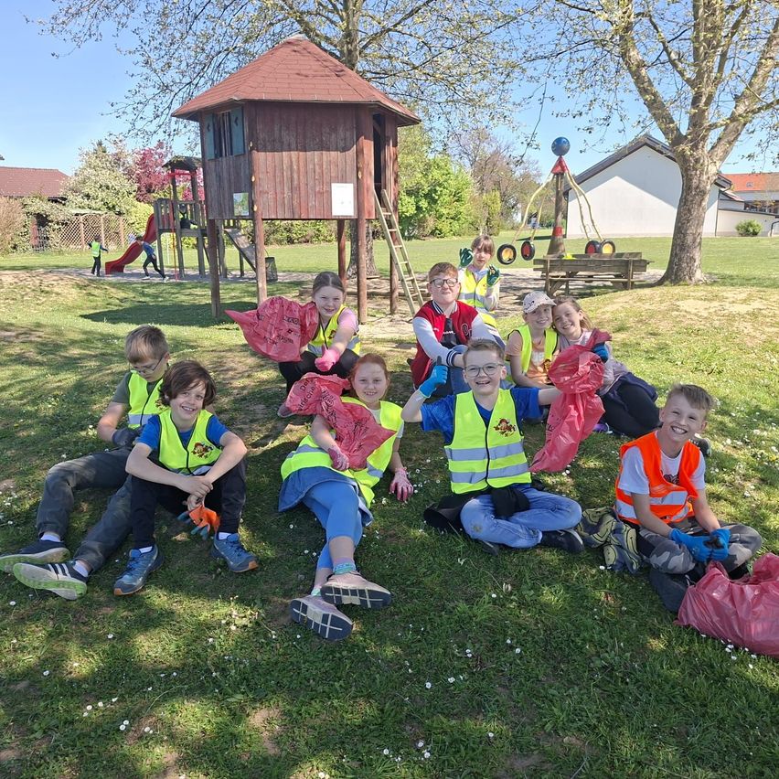 Eine Gruppe von Kindern in Weste sitzt auf Gras und hält Taschen in der Nähe eines Spielplatzes.