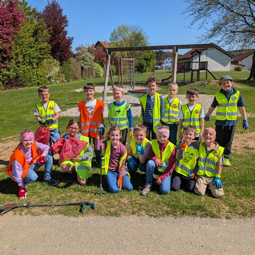 Eine Gruppe von Kindern in leuchtenden Westen posiert für ein Foto in einem Park, hält Mülltüten und einen Besen. Dahinter befindet sich ein Spielplatz mit Schaukeln und ein Haus.