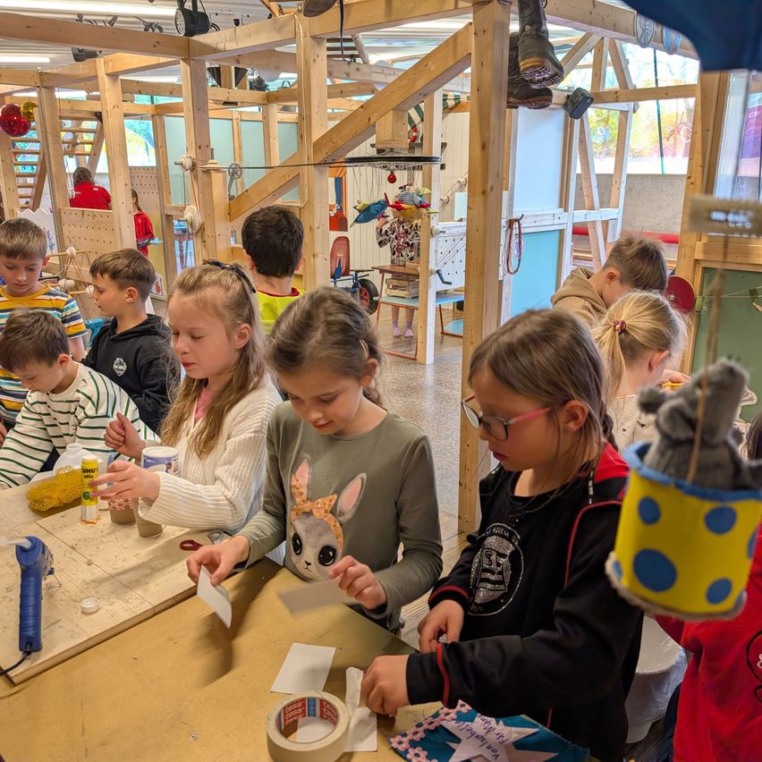 In einem Klassenzimmer sind mehrere Kinder um einen Tisch versammelt und beschäftigen sich mit einer Bastelaktivität. Der Tisch ist mit Materialien wie Kleber, Scheren und Papier übersät. Eine Mädchen in der Mitte hält ein Stück Papier und scheint ihren Mitschülern etwas zu erklären.