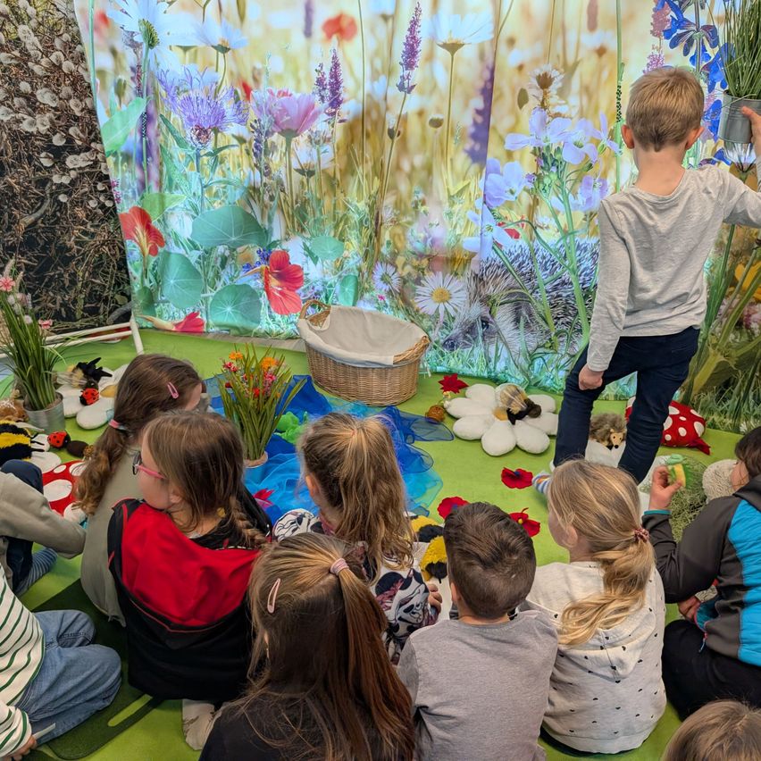 Kinder sitzen auf einem grünen Boden und lauschen einer Geschichte in einem mit Blumentapeten geschmückten Raum. Ein Junge steht mit einem Korb voller Spielzeug.