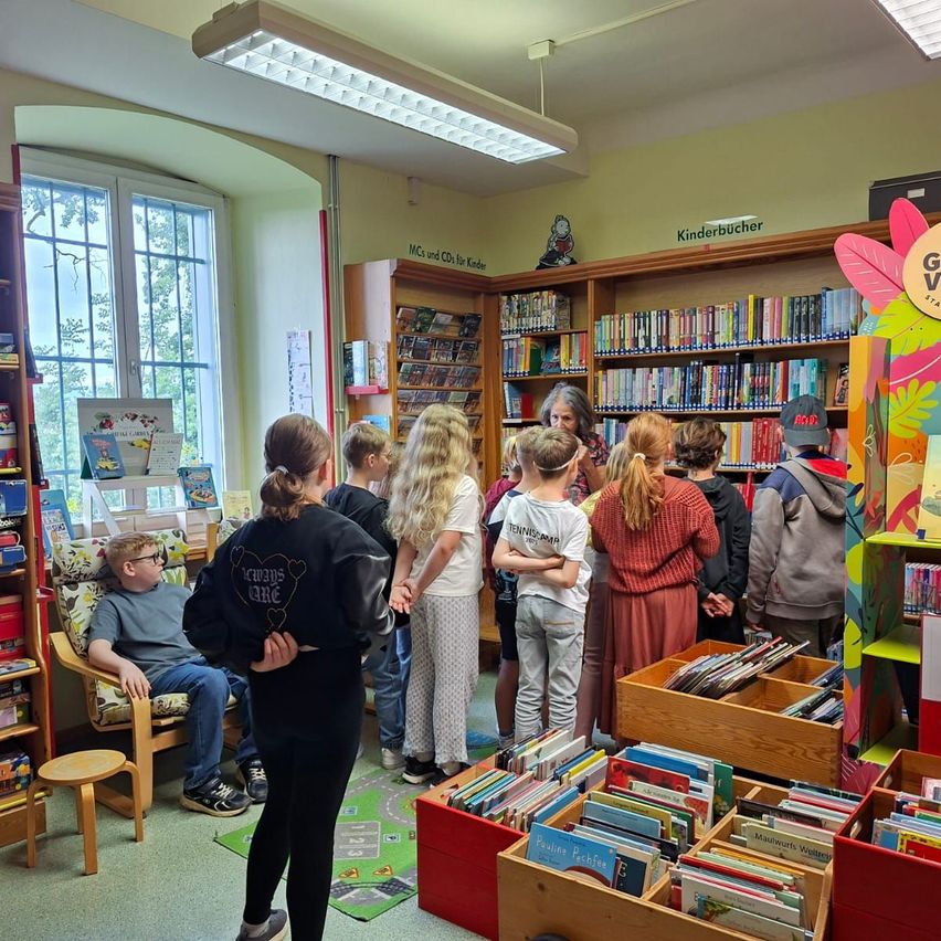Eine Gruppe von Kindern und Erwachsenen in einer Bibliothek, einige stehen, andere sitzen. Der Raum hat hölzerne Regale mit Büchern gefüllt und einen grünen Teppich auf dem Boden. Ein Junge sitzt auf einem Stuhl, und ein Mädchen steht in der Nähe.