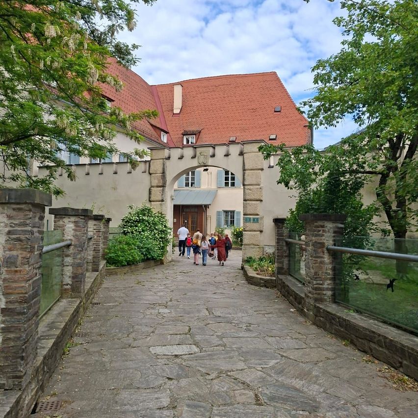 Eine Gruppe von Menschen geht durch den Innenhof eines alten Gebäudes mit rotem Dach. Der Weg ist von Steinsäulen und Grünpflanzen gesäumt.