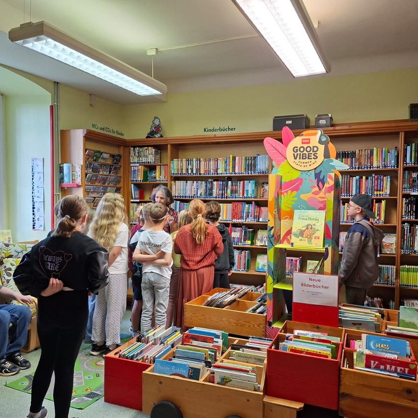 Eine Gruppe von Kindern und Erwachsenen ist in einer Bibliothek und schaut sich Bücher auf farbenfrohen Regalen und Lesetischen an. Ein Schild sagt Gute Stimmung und ein anderes Neue Bilderbücher.