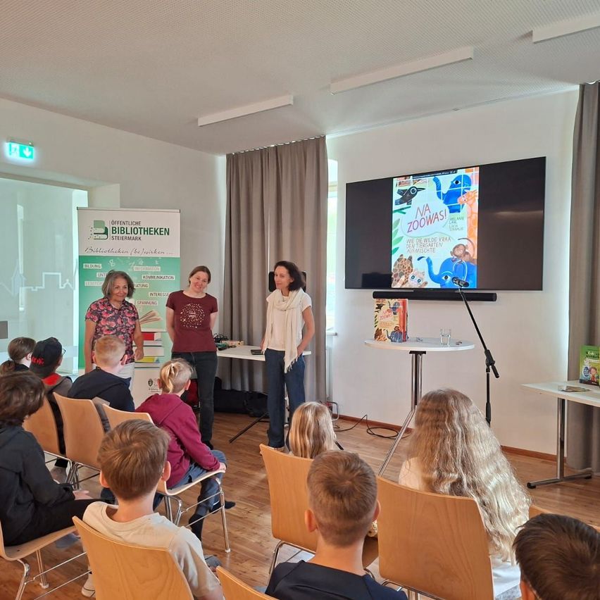 Drei Frauen stehen vor einer Gruppe von Kindern mit Büchern in den Händen. Ein großer Bildschirm zeigt ein farbenfrohes Buch. Der Raum hat Holzböden und beige Stühle.