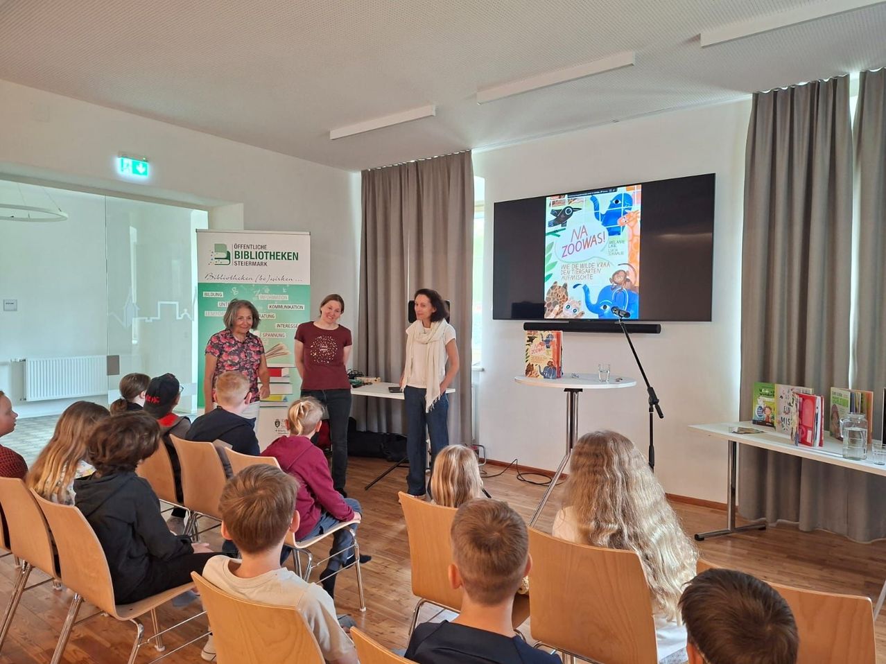Drei Frauen stehen vor einer Gruppe von Kindern mit Büchern in den Händen. Ein großer Bildschirm zeigt ein farbenfrohes Buch. Der Raum hat Holzböden und beige Stühle.