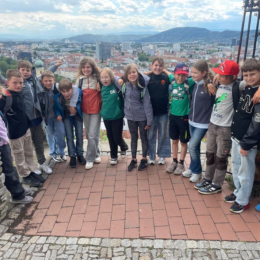 Eine Gruppe von Kindern steht auf einer Ziegelplattform und posiert für ein Foto mit Blick auf die Stadt dahinter. Sie tragen Freizeitkleidung, und einige tragen eine Brille. Sie sehen glücklich und aufgeregt aus.