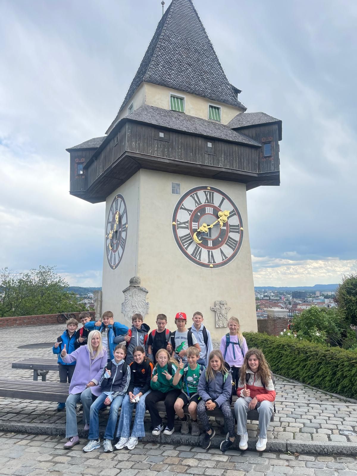 Eine Gruppe von Kindern und Erwachsenen posiert für ein Foto vor einem großen Uhrenturm mit römischen Ziffern.