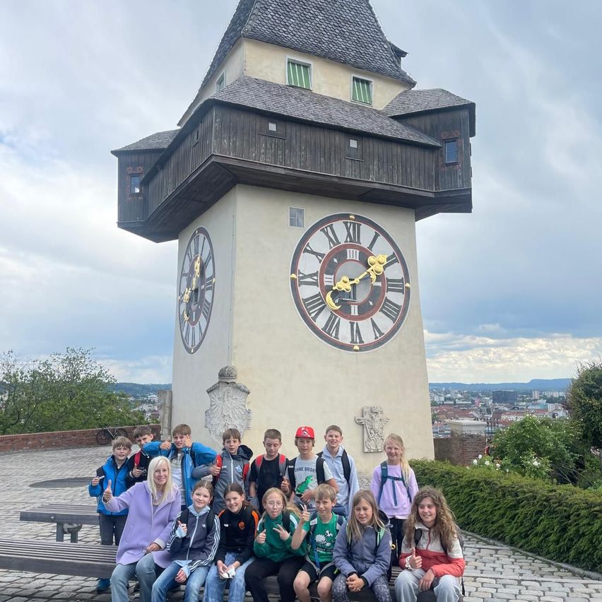 Eine Gruppe von Kindern und Erwachsenen posiert für ein Foto vor einem großen Uhrenturm mit römischen Ziffern.