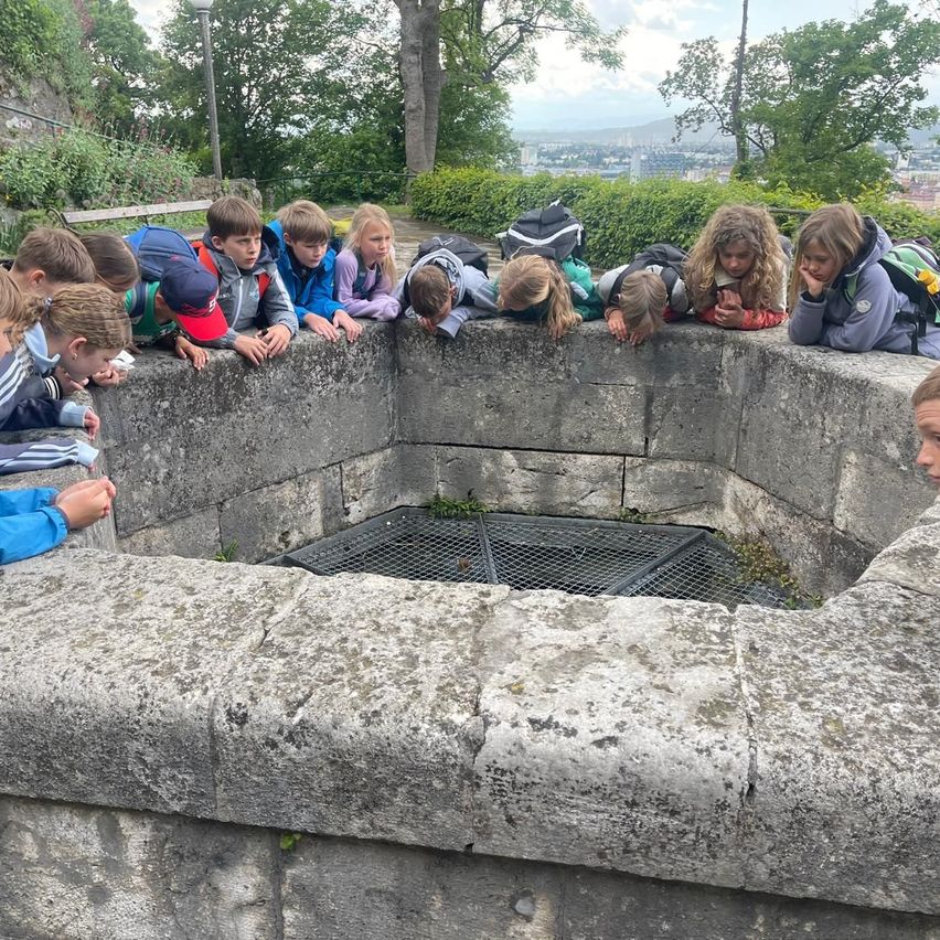 Eine Gruppe von Kindern schaut über eine Steinmauer in einen Brunnen, einige lehnen sich vor, um hineinzusehen. Im Hintergrund sind Bäume und Gebäude zu sehen.