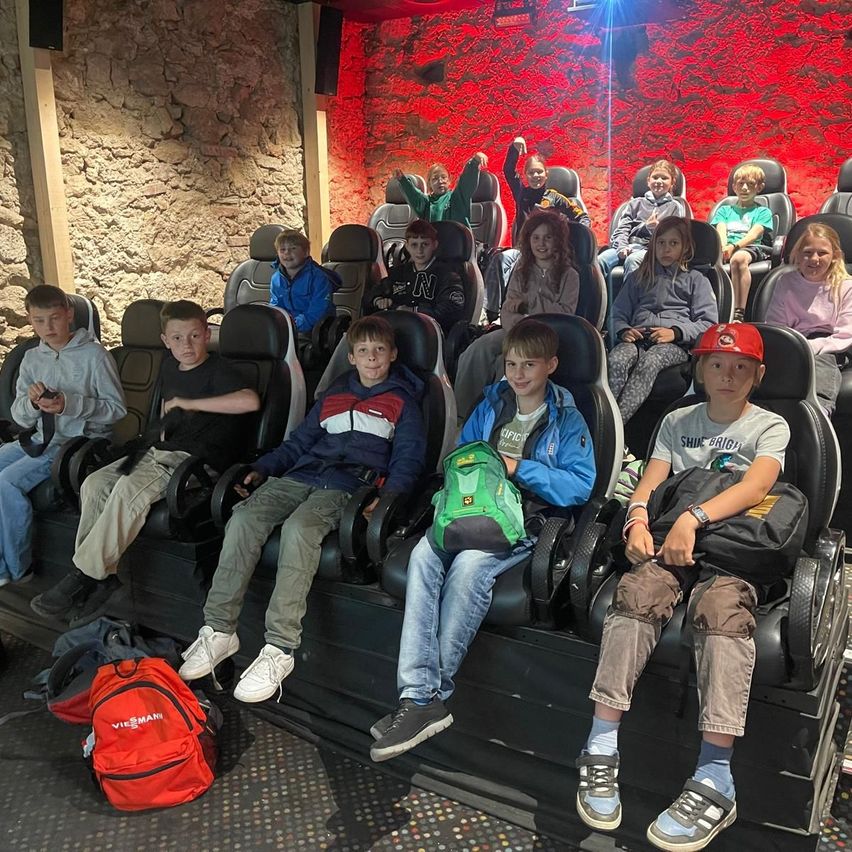 Eine Gruppe von Kindern sitzt in einem Indoor-Kino. Sie tragen Freizeitkleidung und Turnschuhe. Sie haben Rucksäcke auf den Knien. Das Kino hat rote Wände und Sitze.