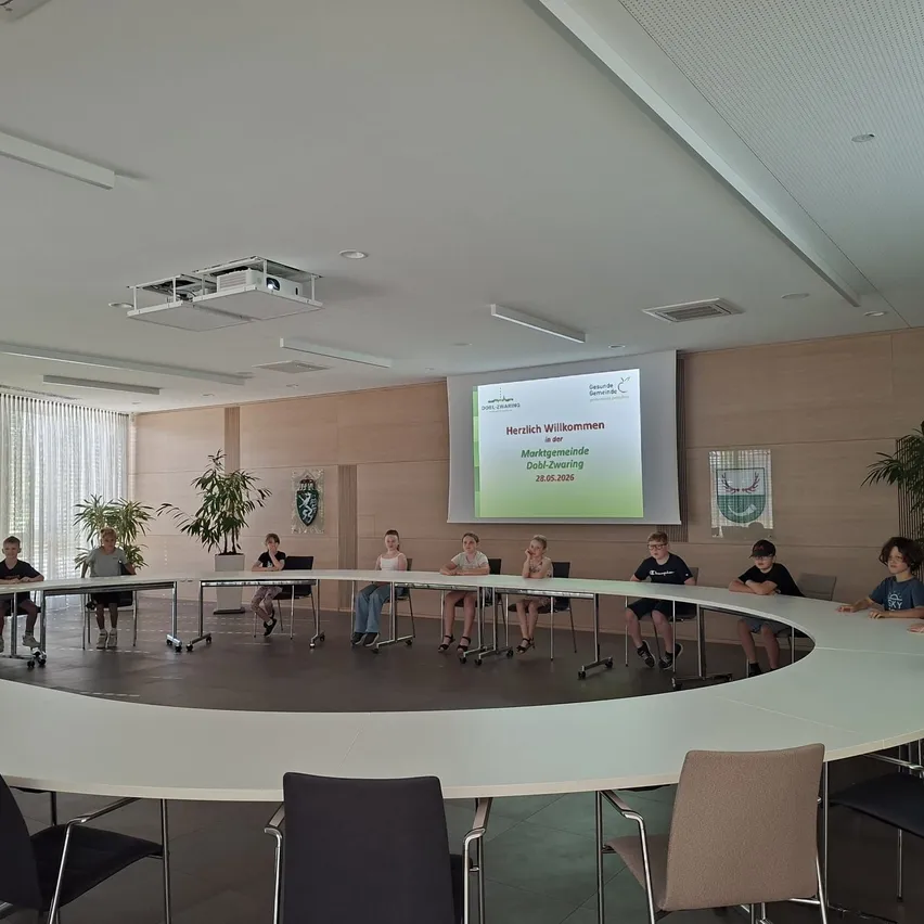 Gruppe von Kindern sitzt um einen Tisch in einem Konferenzraum mit einer Projektionsleinwand, die eine Willkommensnachricht zeigt.