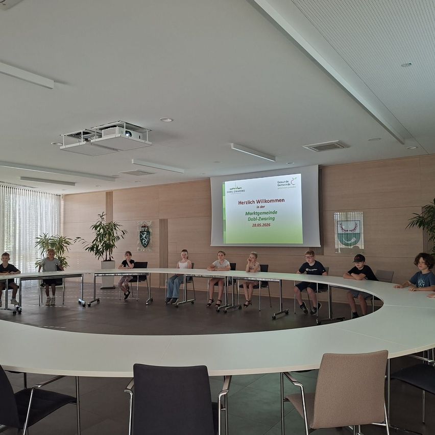 Gruppe von Kindern sitzt um einen Tisch in einem Konferenzraum mit einer Projektionsleinwand, die eine Willkommensnachricht zeigt.