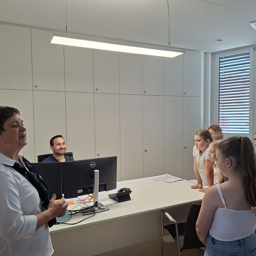 Eine Frau steht vor einem Schreibtisch mit einem Computermonitor und lächelt eine Gruppe von Kindern an, die sich am Schreibtisch lehnen, während ein Mann auf einem Stuhl hinter dem Schreibtisch sitzt.