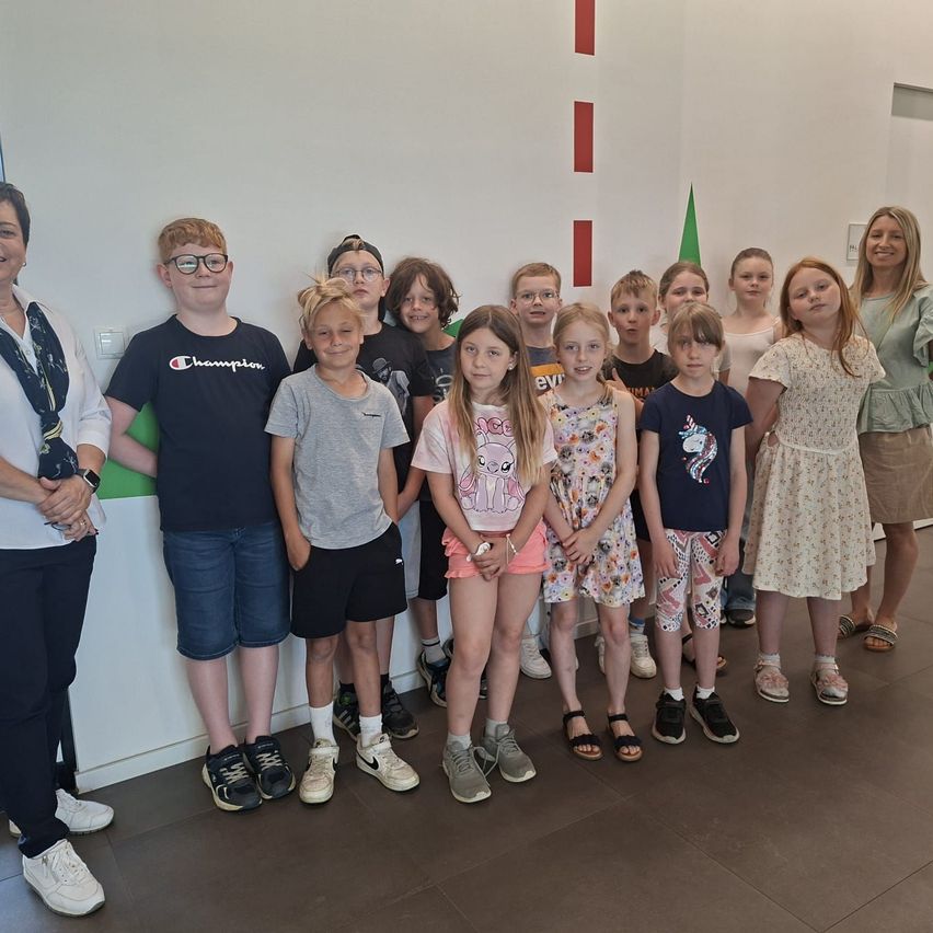 Eine Gruppe junger Kinder und zwei Erwachsene stehen gemeinsam in einem Raum und lächeln für ein Foto. Die Wand hinter ihnen hat rote und grüne Streifen.