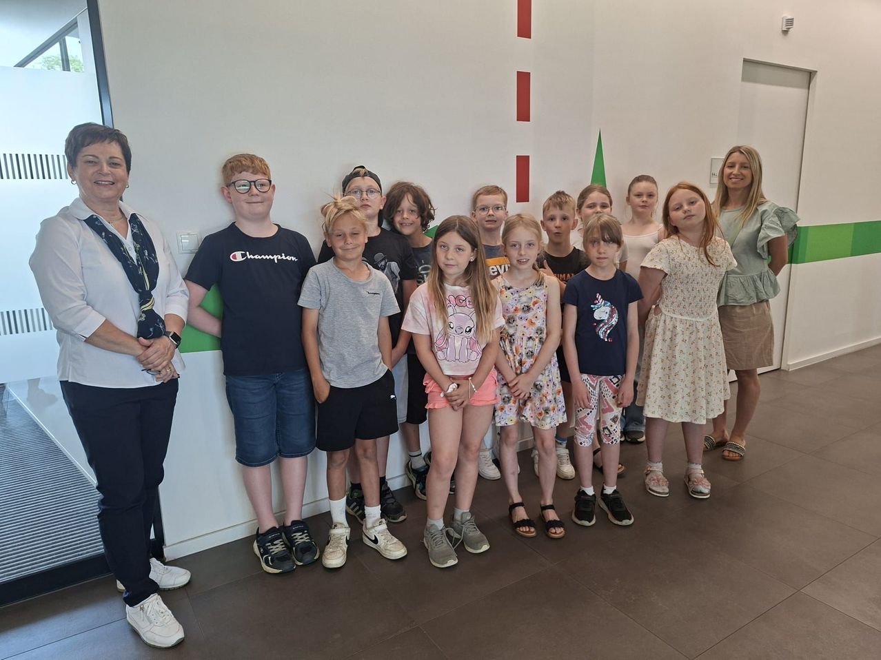 Eine Gruppe junger Kinder und zwei Erwachsene stehen gemeinsam in einem Raum und lächeln für ein Foto. Die Wand hinter ihnen hat rote und grüne Streifen.