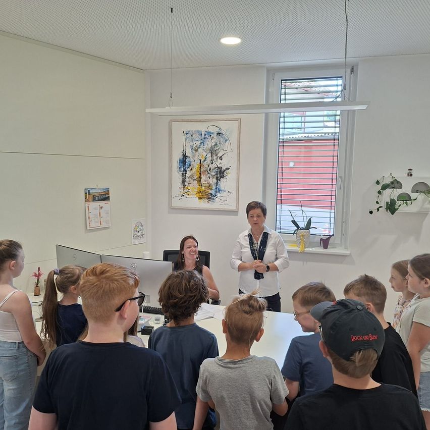 Eine Gruppe von Kindern und zwei Frauen sind in einem Raum. Eine Frau steht vor einem Schreibtisch, die andere sitzt. Ein Monitor und eine Tastatur sind auf dem Schreibtisch. Ein Bilderrahmen und eine Topfpflanze sind an der Wand.