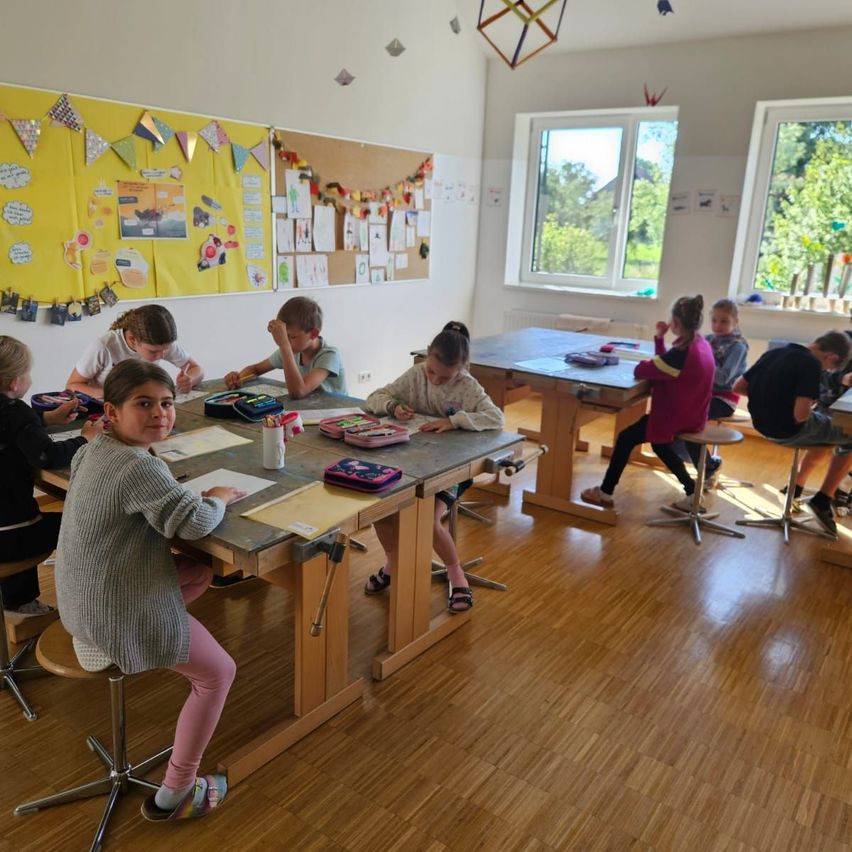 Eine Gruppe von Kindern sitzt um einen Tisch in einem Klassenzimmer herum und arbeitet an ihren Aufgaben. Der Raum ist mit bunten Dekorationen und Postern geschmückt. Sonnenlicht strömt durch die Fenster herein.