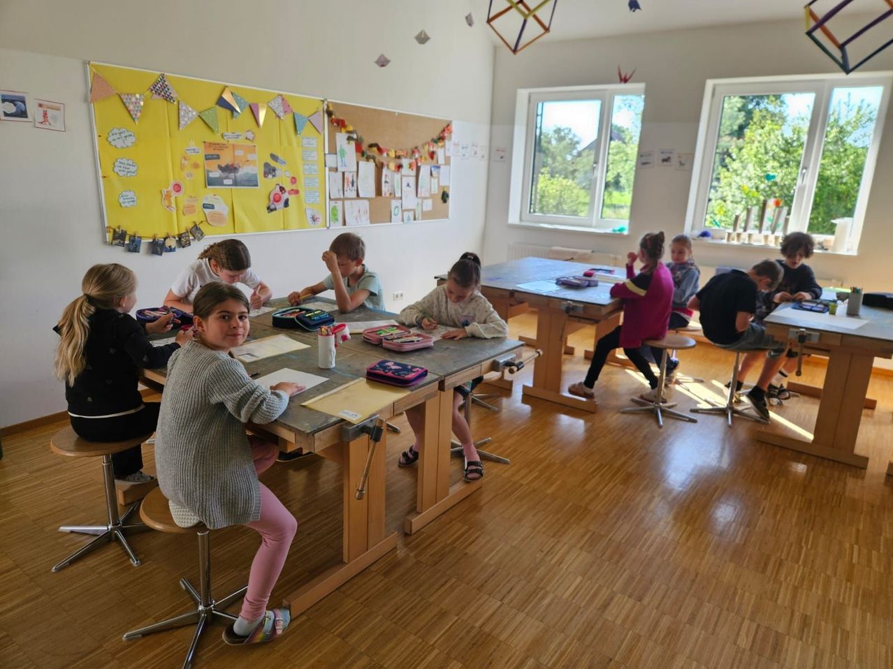 Eine Gruppe von Kindern sitzt um einen Tisch in einem Klassenzimmer herum und arbeitet an ihren Aufgaben. Der Raum ist mit bunten Dekorationen und Postern geschmückt. Sonnenlicht strömt durch die Fenster herein.