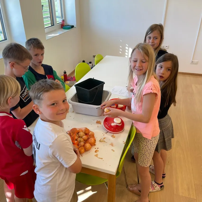 Eine Gruppe von Kindern sitzt um einen Tisch herum und schält Zwiebeln. Ein Mädchen lächelt und hält eine Zwiebel. Hinter ihr blickt ein weiteres Mädchen zu. Auf dem Tisch befinden sich ein Mülleimer und ein Behälter.
