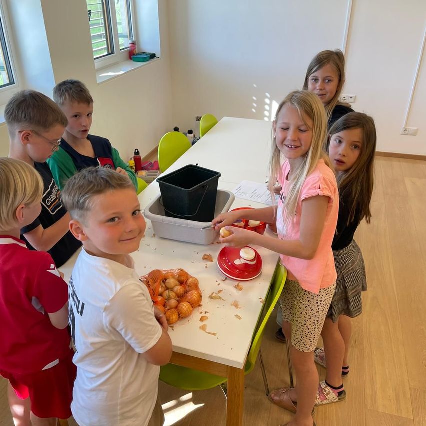 Eine Gruppe von Kindern sitzt um einen Tisch herum und schält Zwiebeln. Ein Mädchen lächelt und hält eine Zwiebel. Hinter ihr blickt ein weiteres Mädchen zu. Auf dem Tisch befinden sich ein Mülleimer und ein Behälter.