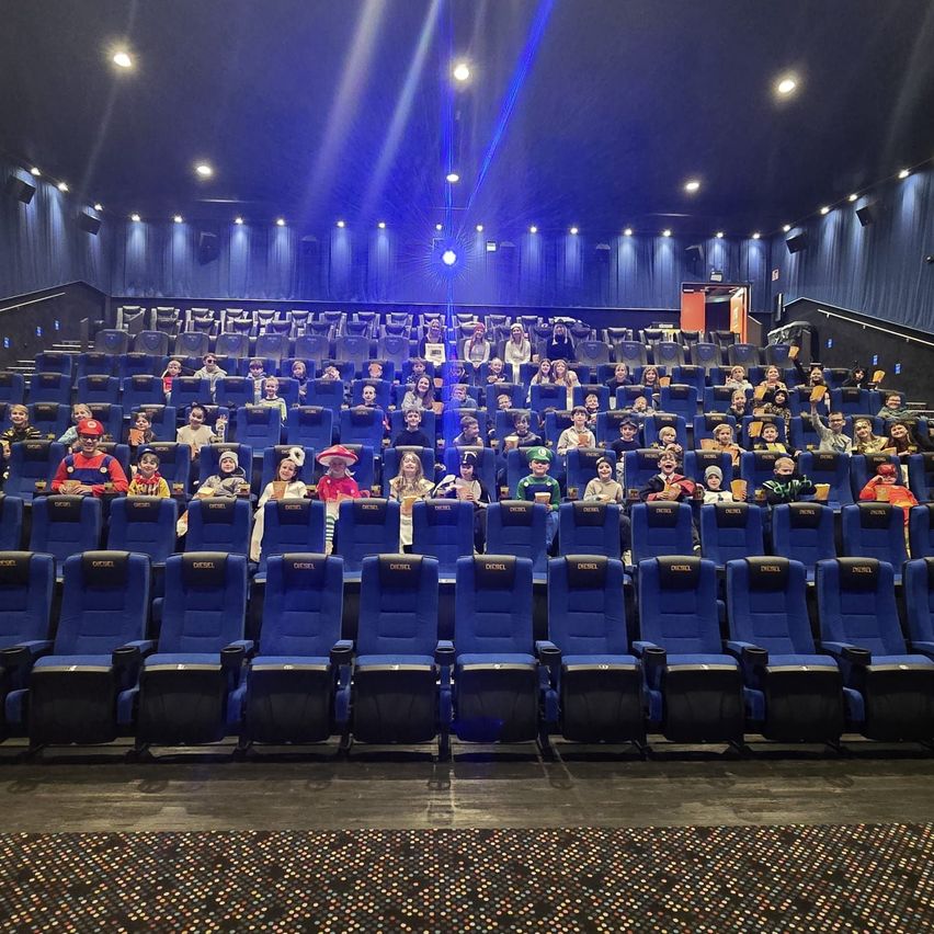 Eine Gruppe von Menschen, wahrscheinlich Kinder, in verschiedenen Kostümen, sitzt in einem Kinosaal mit blauen Stühlen und blauer Beleuchtung.