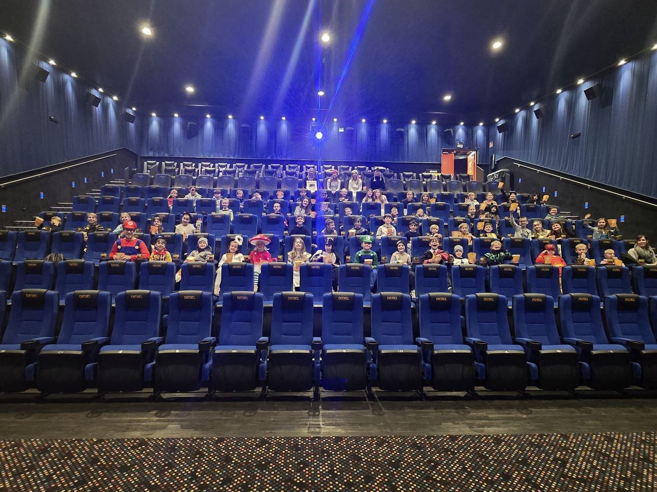 Eine Gruppe von Menschen, wahrscheinlich Kinder, in verschiedenen Kostümen, sitzt in einem Kinosaal mit blauen Stühlen und blauer Beleuchtung.