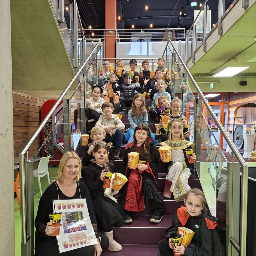 Eine Gruppe von Kindern in Kostümen, die auf Treppen sitzen und Popcorn und Getränke halten, mit einer Frau, die ein Poster vor sich hält.