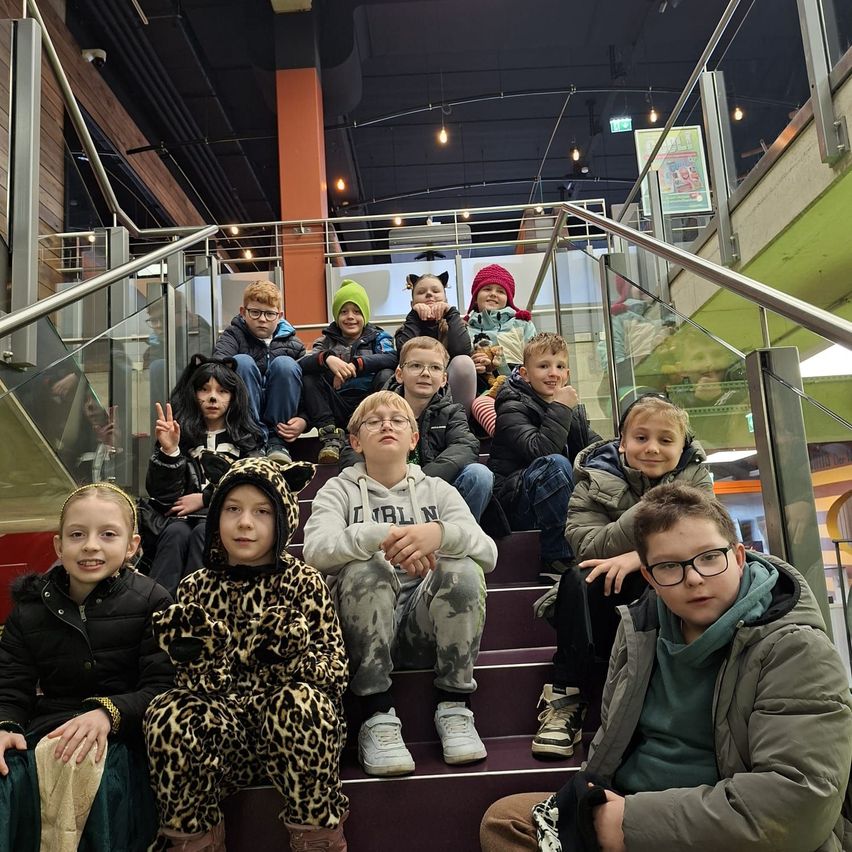 Eine Gruppe von Kindern und Jugendlichen posiert für ein Foto auf einer Treppe. Einige tragen Winterkleidung, andere Kostüme. Die Umgebung scheint im Innenbereich zu sein, mit einem modernen architektonischen Design.