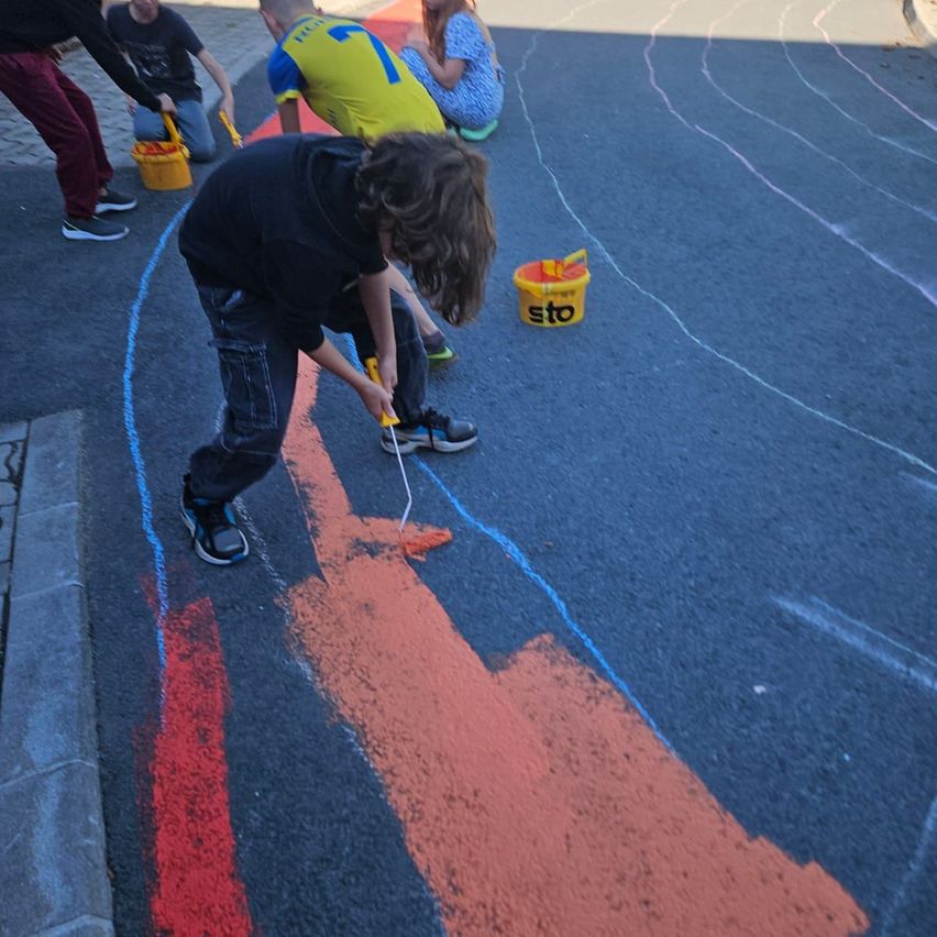 Mehrere Kinder malen ein großes Wandbild auf der Straße mit verschiedenen Farben. Ein Kind benutzt einen Pinsel.