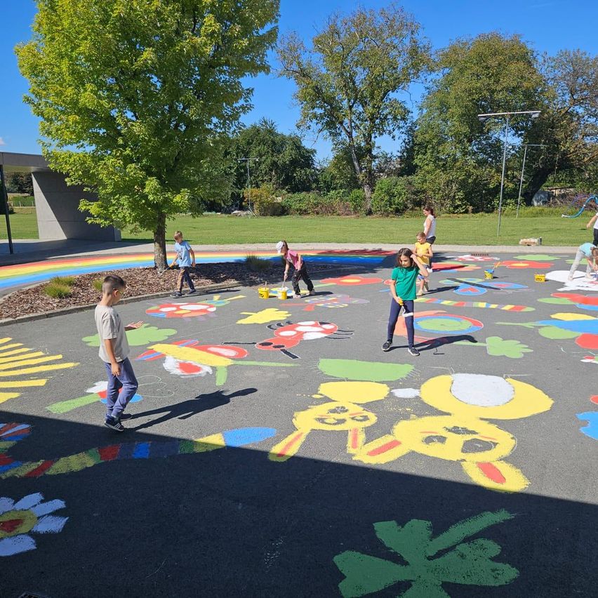 Kinder malen ein großes Wandbild auf dem Boden mit verschiedenen bunten Blumen und Tieren. Sie sind draußen, mit Bäumen und einem Gebäude im Hintergrund.